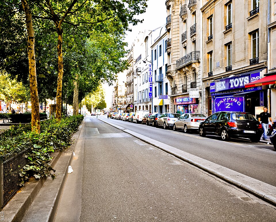 Boulevard de Clichy with sex shop windows, a hotel sign and a pizzeria along one side, cars parked at the kerb and the plane trees of the central walk on the other side.