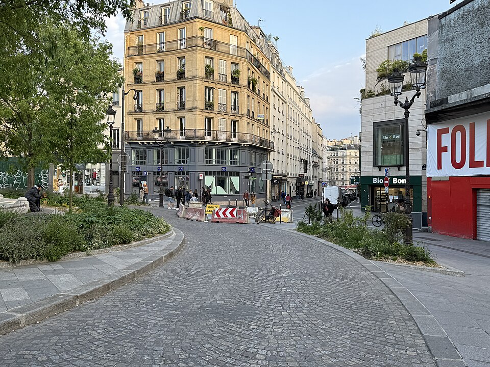 Place Pigalle from the south side, the roadway relaid in setts, a corner apartment building with a bank at street level, and the red front of the Folies Pigalle with its shutters down at the right.