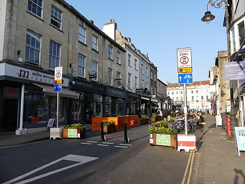 A pedestrianised shopping street of three storey stone terraces, shopfronts at pavement level, flower planters and bollards down the middle of the road.