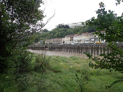 Derelict wooden jetties along the Avon Gorge's tideline, with a run of Georgian and Victorian terraces climbing the wooded hillside behind.