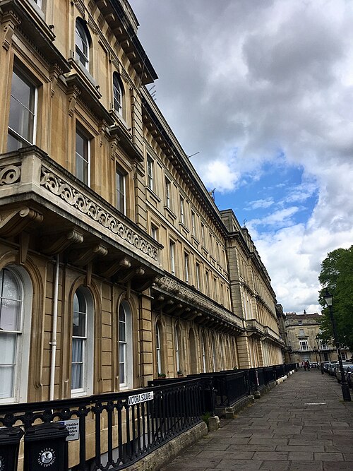 A long unbroken terrace in pale ashlar stone, arched ground floor windows and cast iron balcony railings running the length of the block toward Victoria Square.
