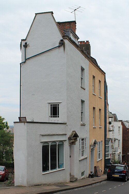A tall terrace on a sloped street, part white render and part ochre, with a stepped gable end, basement railings and cars parked along the kerb.