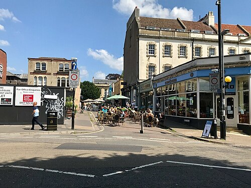 A street corner in Clifton Village, café tables set out on the pavement in front of white rendered shopfronts.