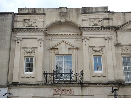 A carved limestone facade with paired pilasters, a pedimented window and a wrought iron balcony rail, part of a converted hotel building now used as shops.