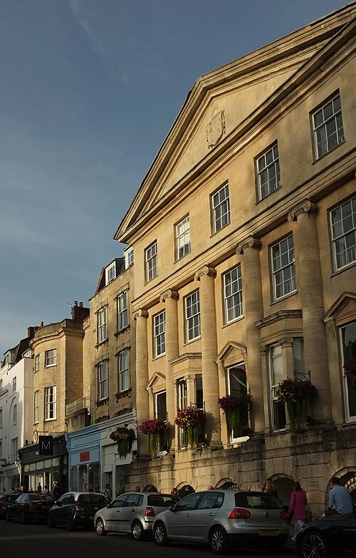 A row of Regency stone buildings along The Mall, ionic columns and a pediment over a corner entrance, cars parked along the kerb and flower boxes at the shopfronts.