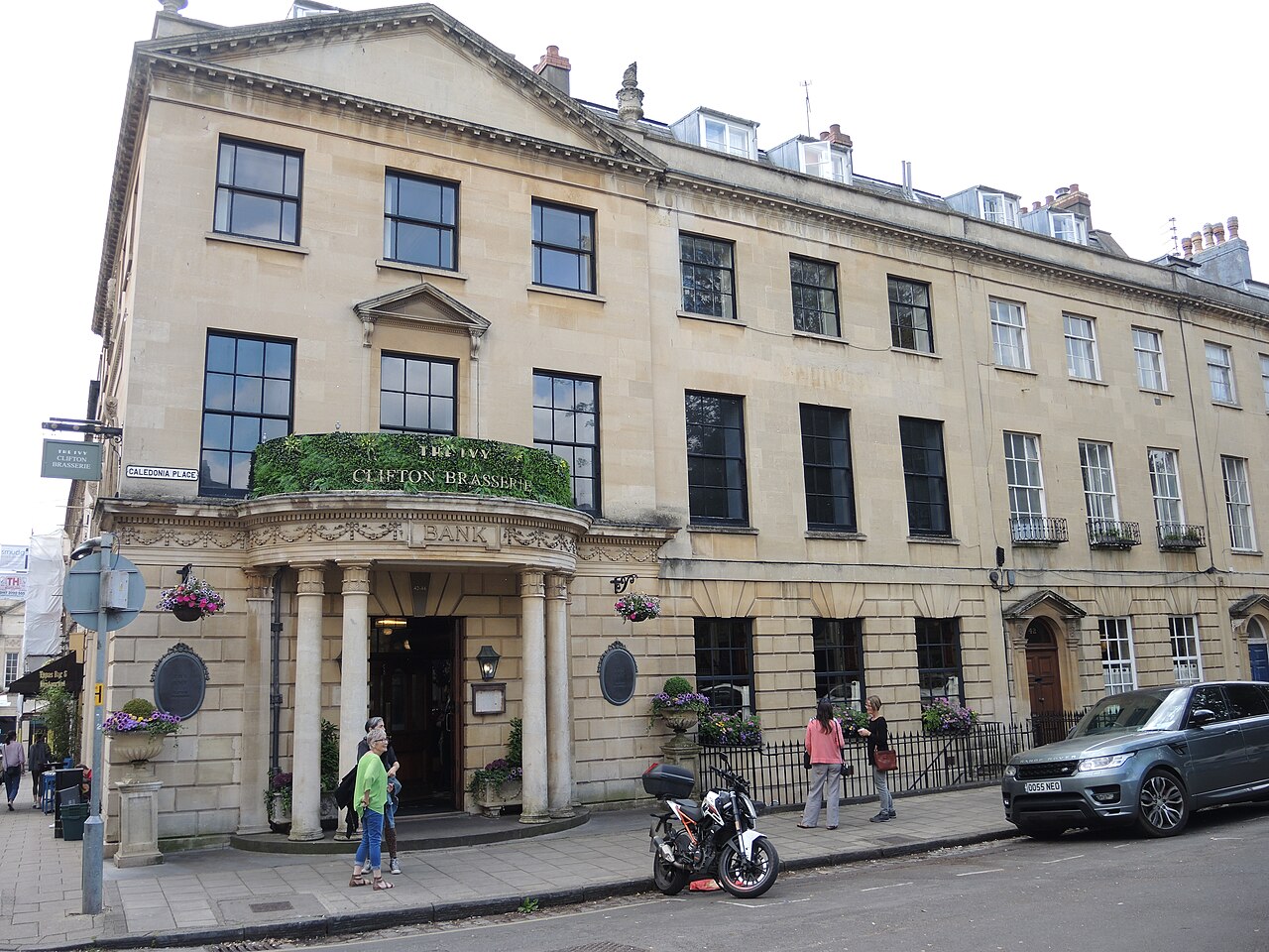 A columned corner building at Caledonia Place, a curved portico projecting over the pavement with BANK carved into the stonework above it, and a run of arched ground floor windows continuing the block along the street.