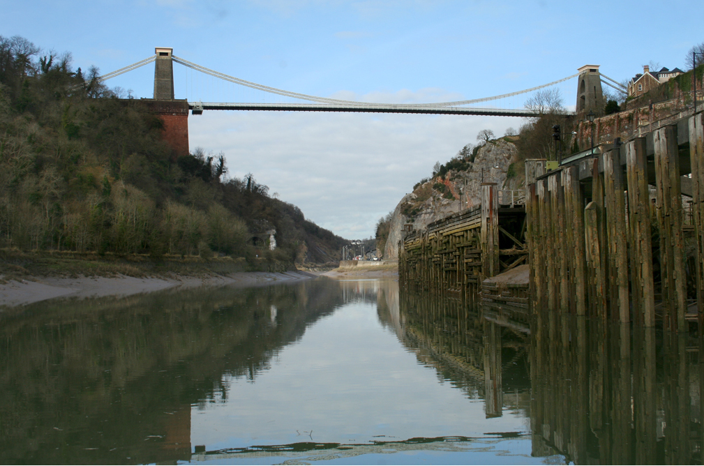 The suspension bridge crossing high above the Avon Gorge on two stone towers, wooded cliffs on both sides and derelict timber jetties on the riverbank below.