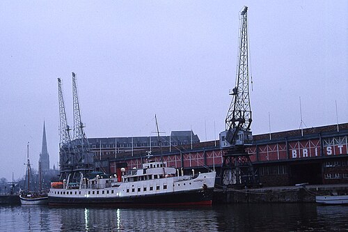 An older photograph of the Balmoral moored beside the electric cranes and the dockside museum shed, a church spire behind.