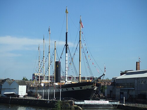 The SS Great Britain in dry dock, masts hung with signal flags, dockyard sheds and a brick chimney behind.