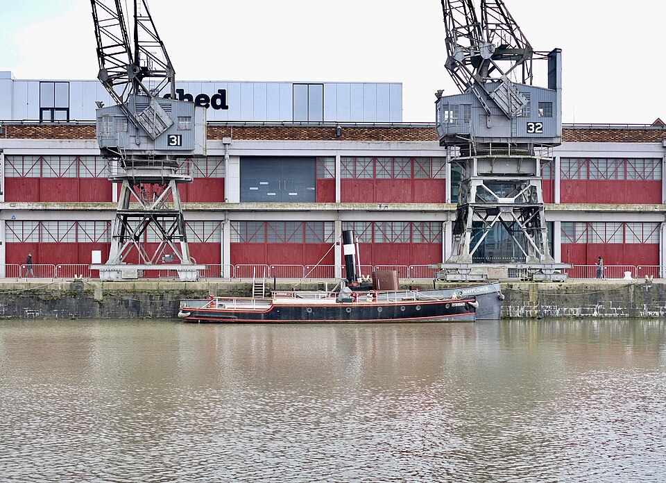 Cranes 31 and 32 in front of the M Shed's red and grey transit shed wall, the tug Mayflower moored at the foot of the quay.
