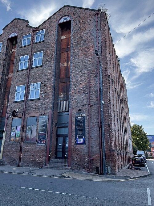 The corner warehouse on Blundell Street, the date 1916 set in the arch over a tall boarded loading bay and 1882 on the gable beside it, red painted downpipes and a long blank flank wall running back down the street.