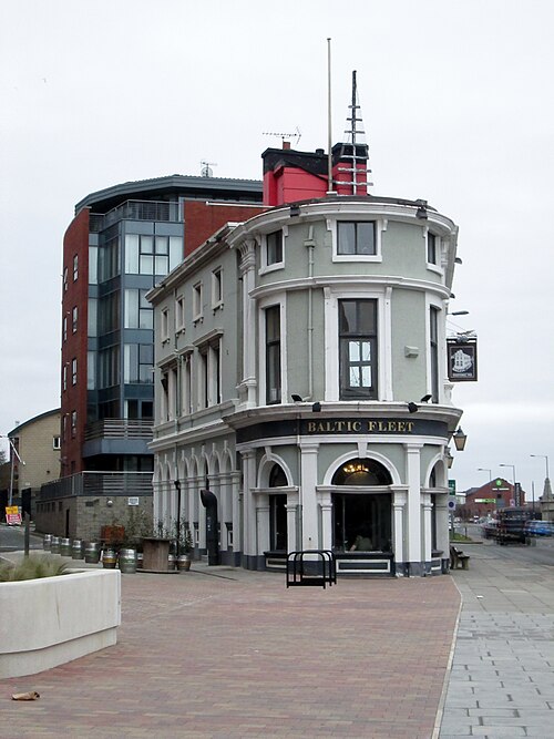 The Baltic Fleet on Wapping, a flat iron pub with a curved end, arched windows at street level and a ship's mast and rigging on the roof, with a newer flats block behind it.