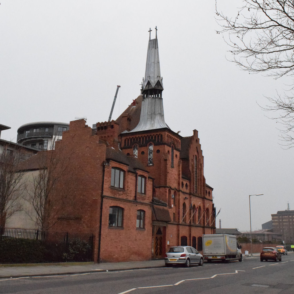 Gustav Adolf Church on Park Lane, red brick with a lead covered spire and two crosses, cars and a box van at the kerb below it.