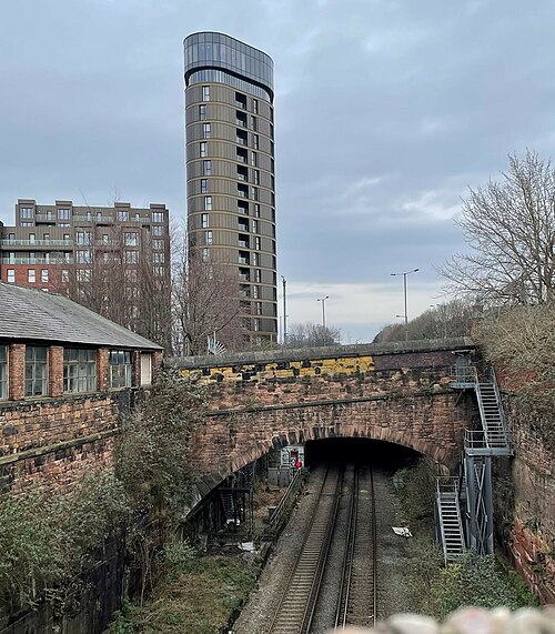 The cutting at the old St James station, a sandstone bridge over the tracks with steel access stairs down to track level, a small brick workshop with steel windows on the left and a round clad tower behind.