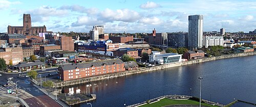 The district from the air, looking inland across the dock. Brick housing along the quay, blue clad sheds and warehouses behind it, newer towers on the right and a cathedral on the ridge.