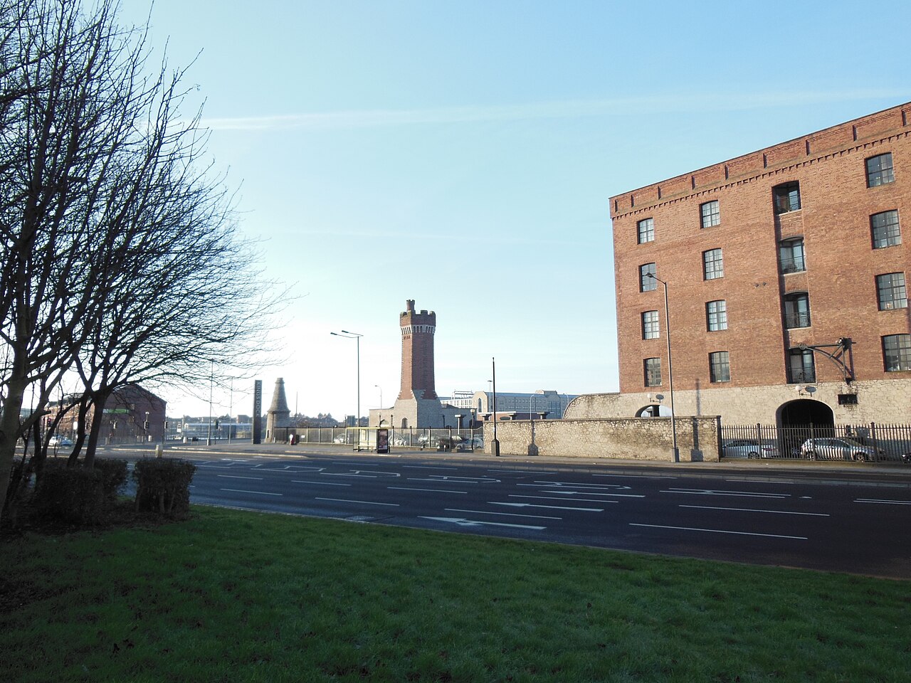 The wide carriageway on Wapping on a clear winter afternoon, the castellated brick hydraulic tower and the stone dock wall on one side, the arched brick warehouse at Wapping Dock on the other.