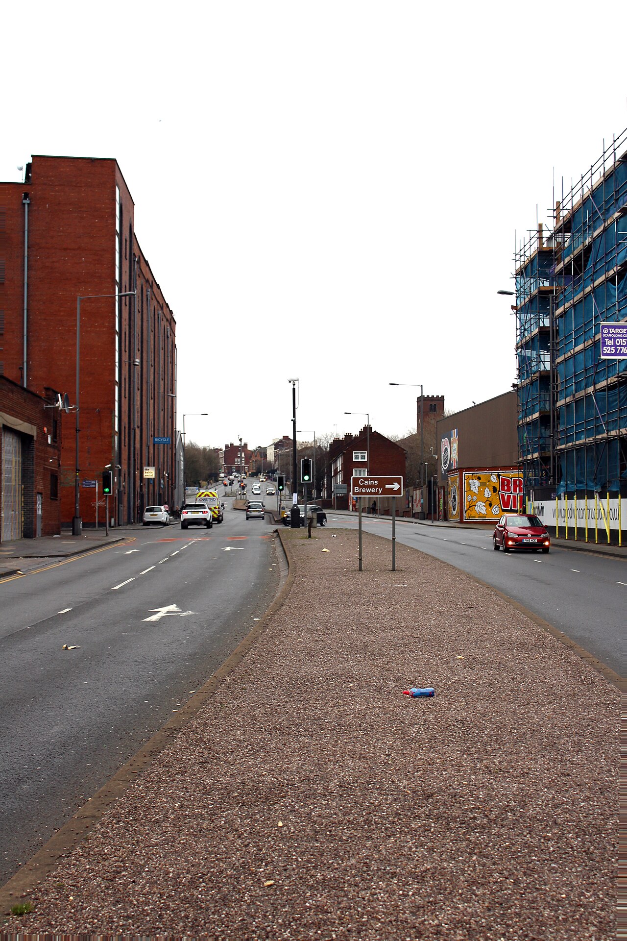 Parliament Street from the middle of the road, a long brick warehouse along the left, a building under scaffolding and sheeting on the right, and a brown sign pointing to Cains Brewery.