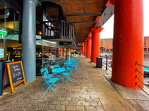 The colonnade at quay level, cast iron Doric columns painted red carrying the brick arcade above, cobbles underfoot and a cafe's tables set out beneath it.