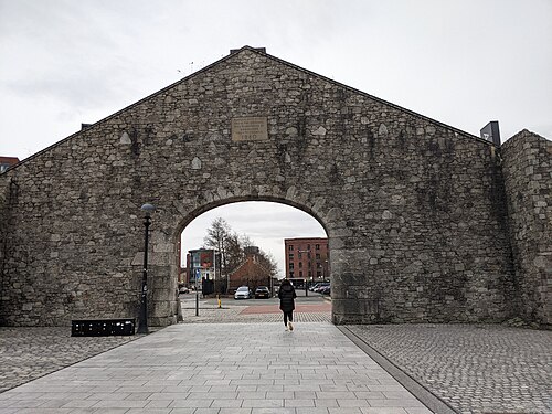 A stone arch in the dock's boundary wall, rebuilt in 1980 from an earlier warehouse on the site, with a dedication plaque set into the gable above the opening.