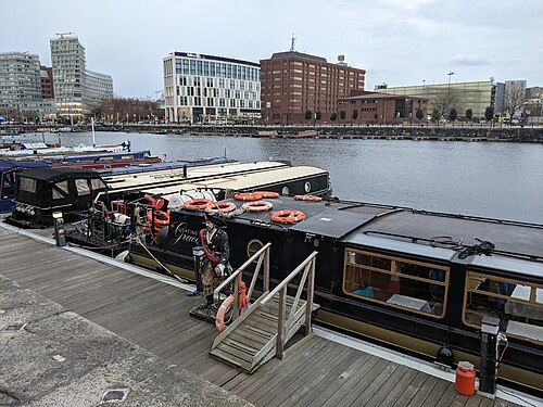 Narrowboats moored close along the quay, fenders and mooring lines between them, modern office and hotel buildings visible on the far side of the water.