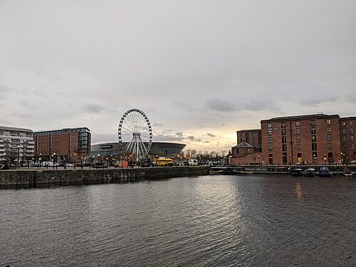 The Albert Dock and Atlantic Pavilion warehouse lettered on its gable at dusk, narrowboats along the quay below it and bare trees strung with lights on the promenade.