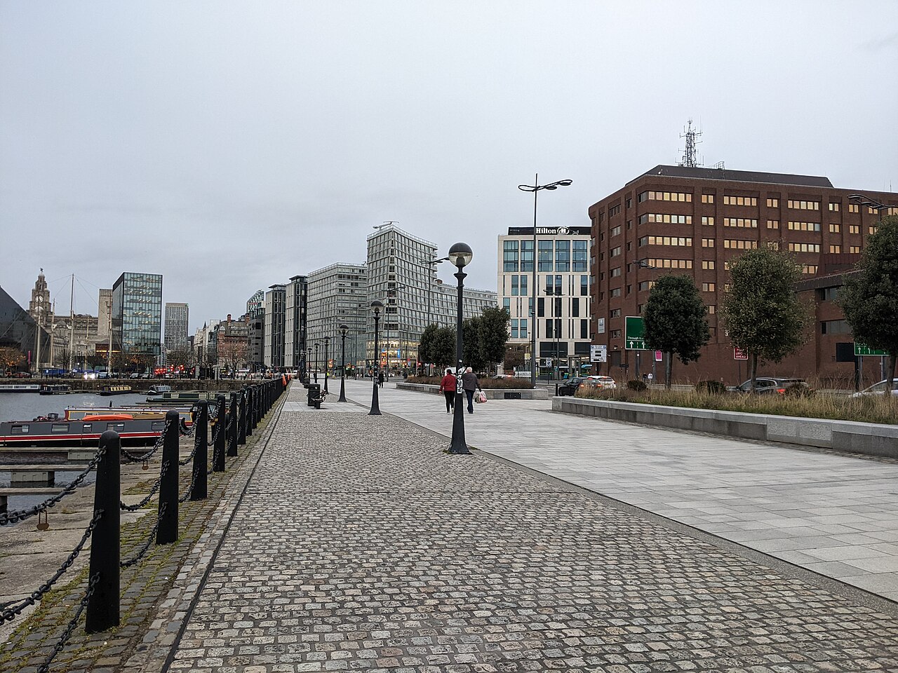 The granite quayside running along the west side of the dock, narrowboats moored two deep against the wall, warehouse blocks and the Liverpool skyline beyond the water.