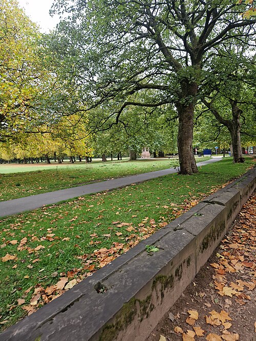 Open meadow crossed by a tarmac path under mature trees, a stone memorial visible in the middle distance.