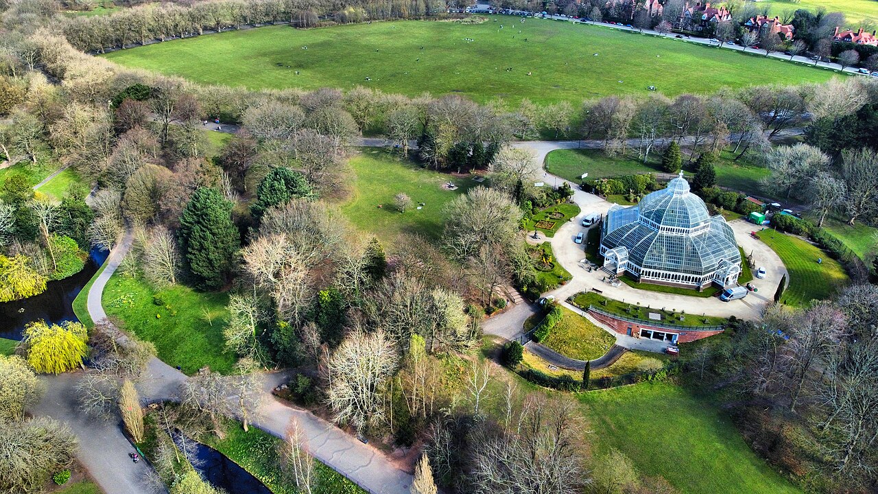 An aerial view over Sefton Park, open meadow behind, the Palm House's glass dome at centre with paths looping around it, and the tree lined lake curving away below.