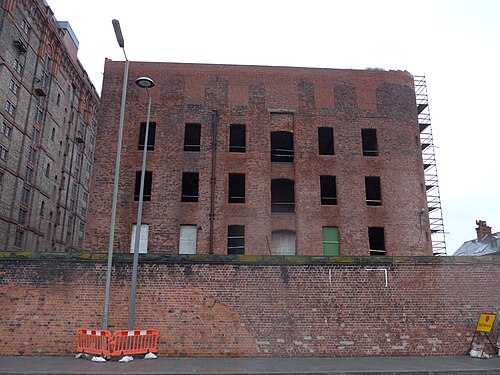 The derelict Stanley Warehouse south of the Tobacco Warehouse, its window openings boarded or left open, scaffolding up one corner and a street lamp and traffic barrier on the pavement below.