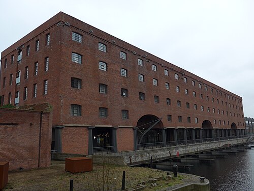 The former north warehouse, now the Titanic Hotel, five storeys with round arched window openings, a colonnade of columns at ground level glazed in behind them, and a pontoon walkway with mooring berths running out over the water.