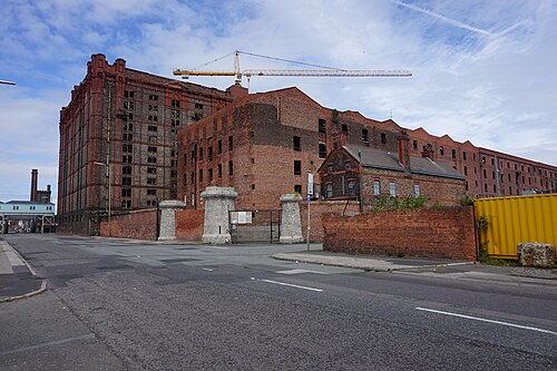Regent Road at the dock's entrance, stone gate piers and a small round windowed lodge building beside the gates, the Tobacco Warehouse's upper floors visible behind the wall, and a crane over the roofline.