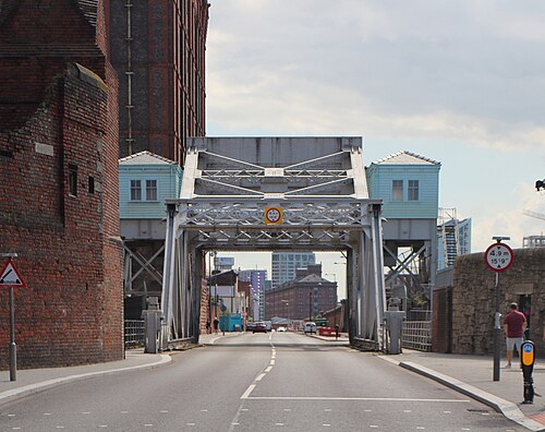 The bascule bridge over Regent Road at the dock's western entrance, a riveted steel lattice span between two pale blue counterweight houses, with a height limit sign and the Tobacco Warehouse's corner visible at left.