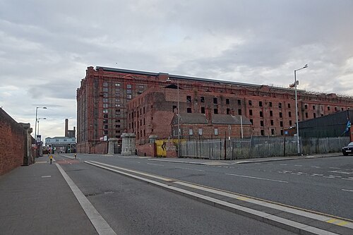 Regent Road at the corner of the dock, the Tobacco Warehouse's grid facade above and the derelict Stanley Warehouse beside it with broken window openings, a smaller brick building with a chimney at the corner, and a palisade fence around the vacant yard.