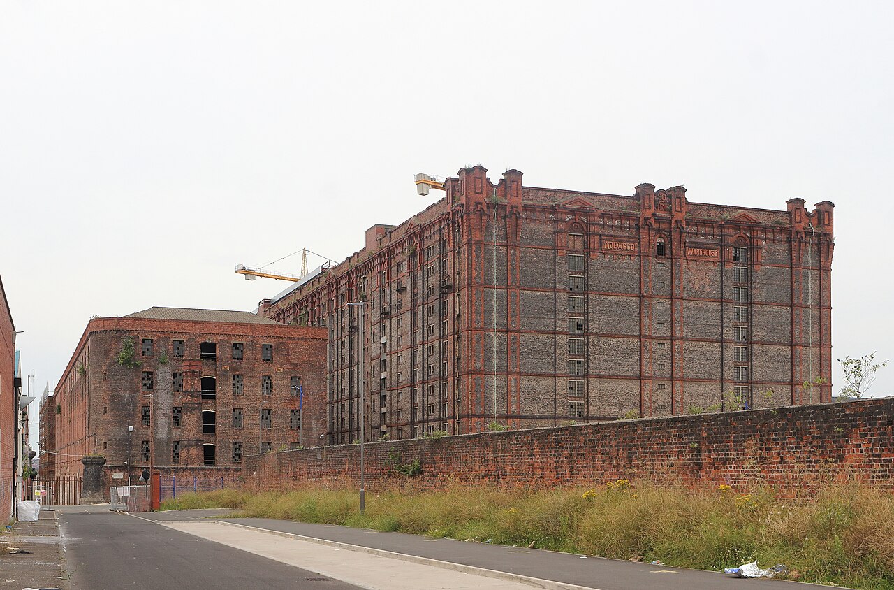 A paved road running toward the Stanley Warehouse and the Tobacco Warehouse, the derelict Stanley Warehouse in the foreground and the Tobacco Warehouse's full fourteen storey height behind it, TOBACCO WAREHOUSE lettering across its cornice and a crane over the roofline.