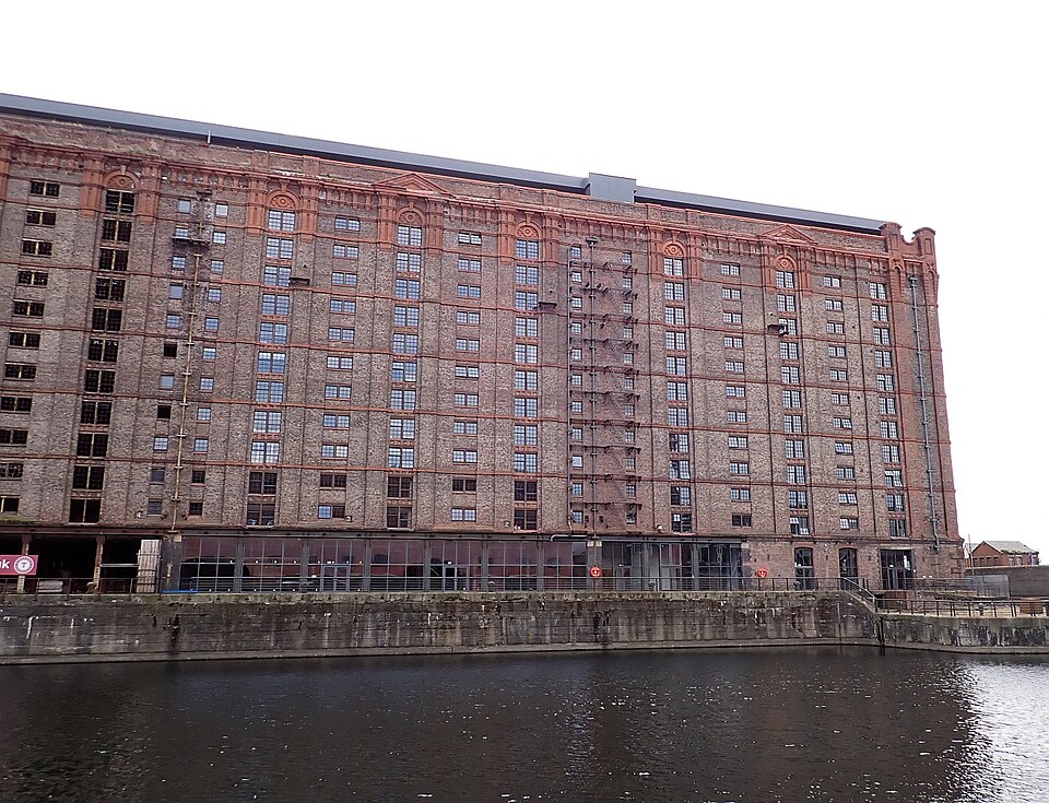 The Tobacco Warehouse's west end across the dock basin, its full height of window bays reflected in the water, a glazed arcade and walkway along the ground floor behind slim black piers, and mooring pontoons in the foreground.