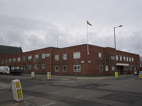 A two storey red brick police station on a street corner, a Union flag on its pole and marked patrol vehicles at the kerb.