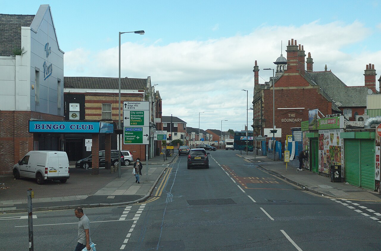 A commercial street with low rise shopfronts, a bingo club and a corner pub called the Boundary, overhead wires and directional road signs.