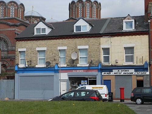 Two storey pale brick shops with dormer windows, a newsagent and an Afro-West Indian grocer at street level, the Greek Orthodox church's domes visible behind the roofline.