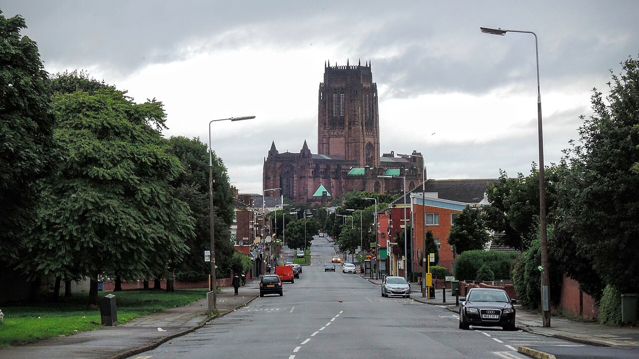 A straight street lined with trees and brick terraces climbing toward Liverpool's Anglican Cathedral, its sandstone tower filling the skyline at the far end.