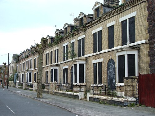 A row of yellow brick terraced houses with dormer gables and stepped parapets, upper and lower windows boarded, ironwork railings along the pavement.