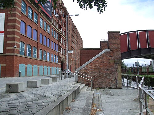 The Royal Mills corner at Henry Street, arched windows with blue and teal panels under the sills, a red iron footbridge crossing the Rochdale Canal on the right.