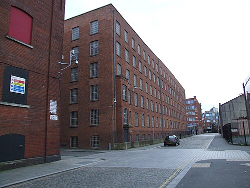 A mill block running the length of Jersey Street, rows of barred windows in dark brick receding to a taller building at the far end, a single car parked at the kerb.