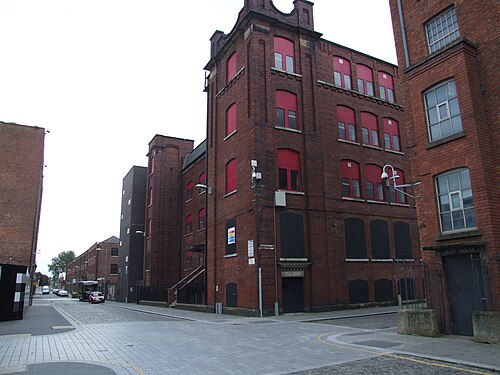 A mill building on a street corner in ochre and red brick, red blinds behind arched window frames and a stone pediment at the roofline.