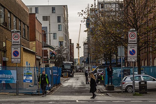 A street under redevelopment near Bengal Street, blue hoarding and a crane behind bare trees, a new apartment block and an older mill front on either side.