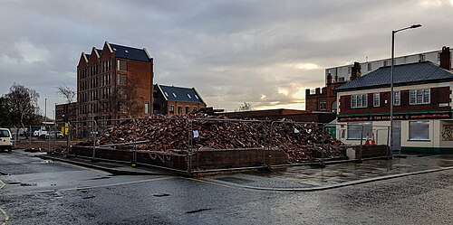 A cleared corner site behind steel fencing, brick rubble piled inside it, the Shamrock pub and a new brick apartment block standing on either side.