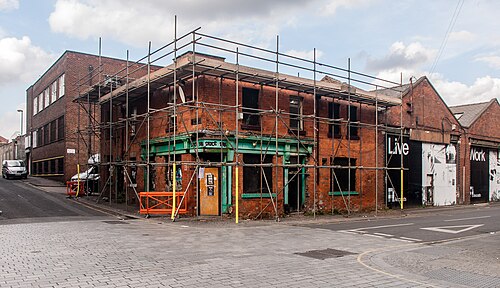 A red brick corner building behind scaffolding, a green painted former pub front at street level and LIVE WORK hoarding covering the mill wall beside it.
