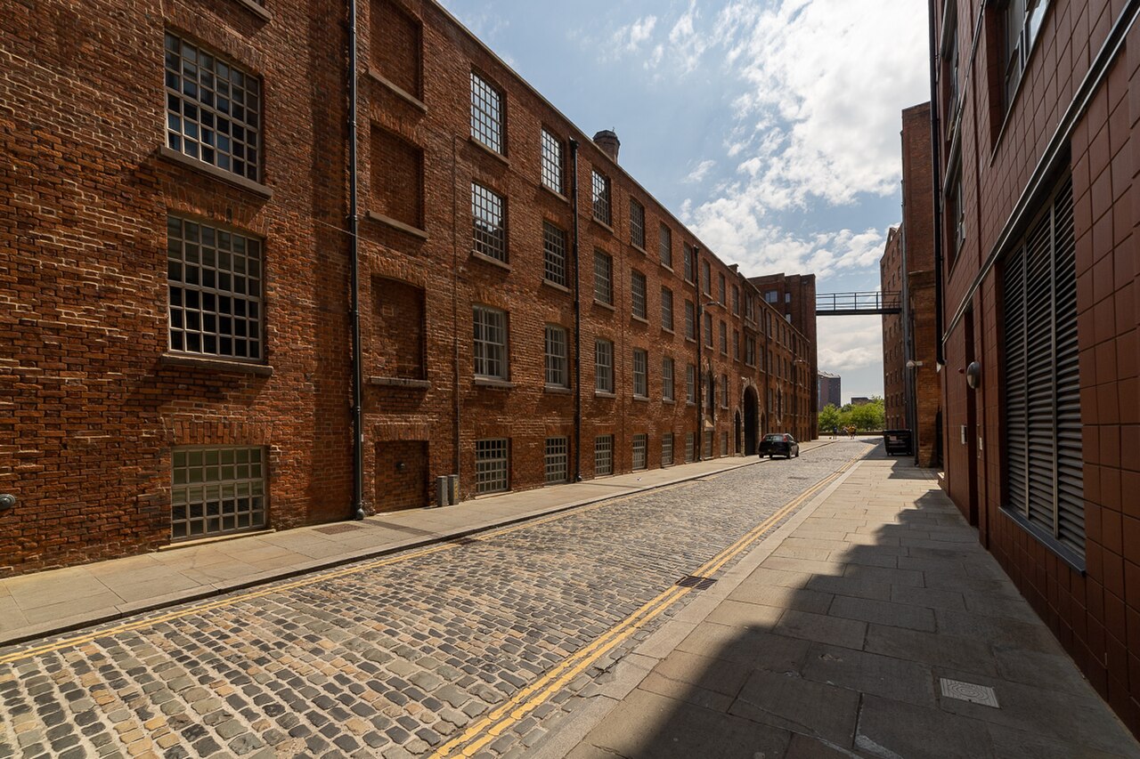 A cobbled lane between the Old Mill, Decker Mill and New Mill buildings on Murray Street, tall brick mill walls with small paned windows on both sides and a covered iron walkway crossing overhead.