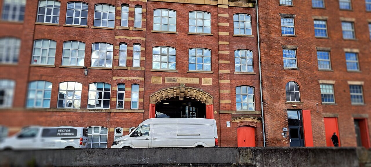 The brick archway into Royal Mill on Redhill Street, ROYAL MILL cut into the stone lintel above the opening, red painted doors along the ground floor and a canal wall in the foreground.