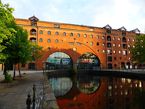 Middle Warehouse's round brick arches and glass infill reflected in the still canal water, a cobbled towpath and trees in front.