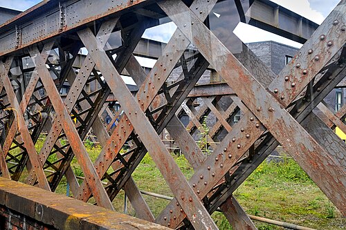 Riveted steel lattice girders of a railway bridge, streaked with rust, seen close against a brick parapet.